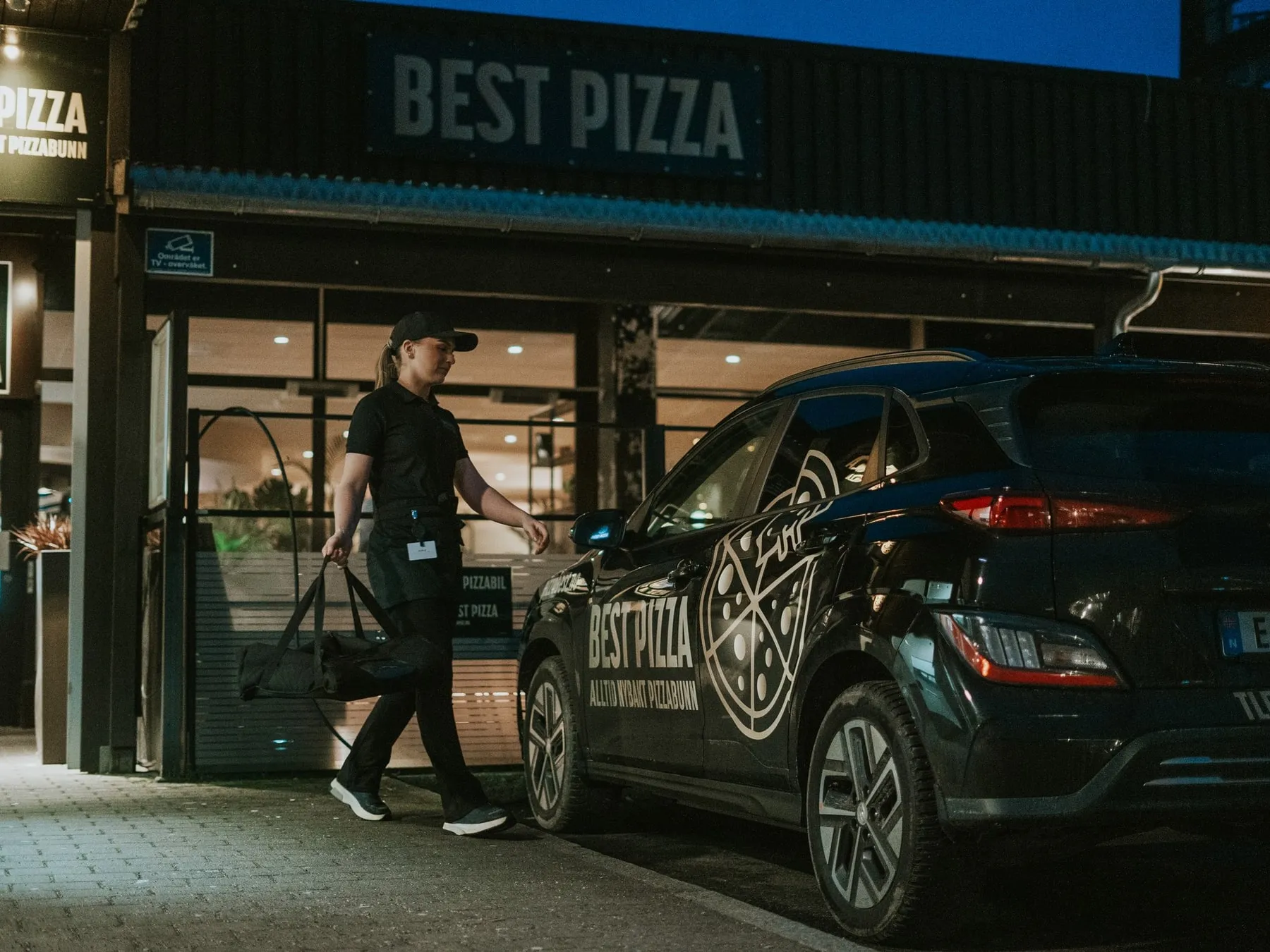 A Best Pizza staff member carrying the delivery bag to the branded car outside the restaurant at dusk.